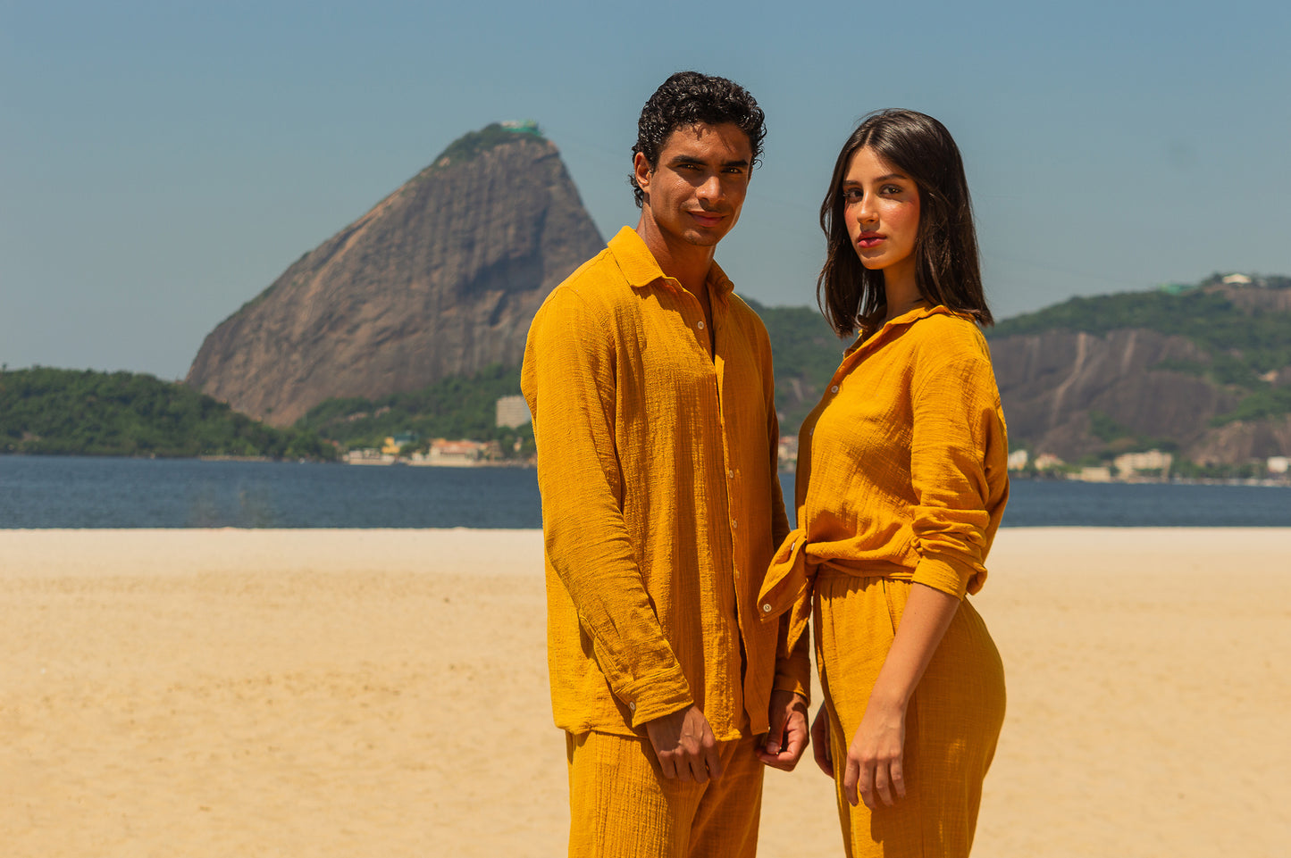 Casal vestindo full look com camisa manga longa e calça amarelo dijon unissex, posando de frente em ambiente de praia.
