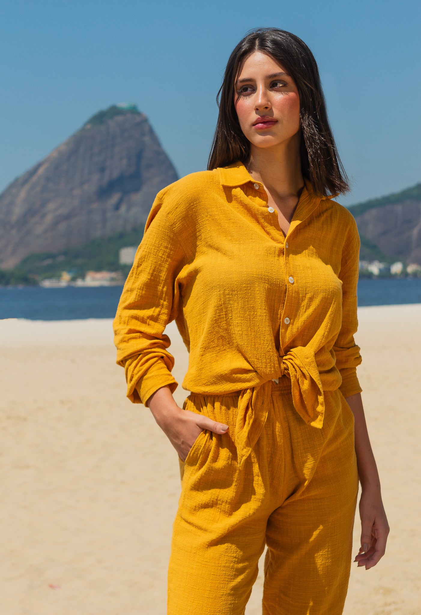 Modelo feminina vestindo full look com camisa manga longa e calça amarelo dijon unissex, em pose frontal na praia.