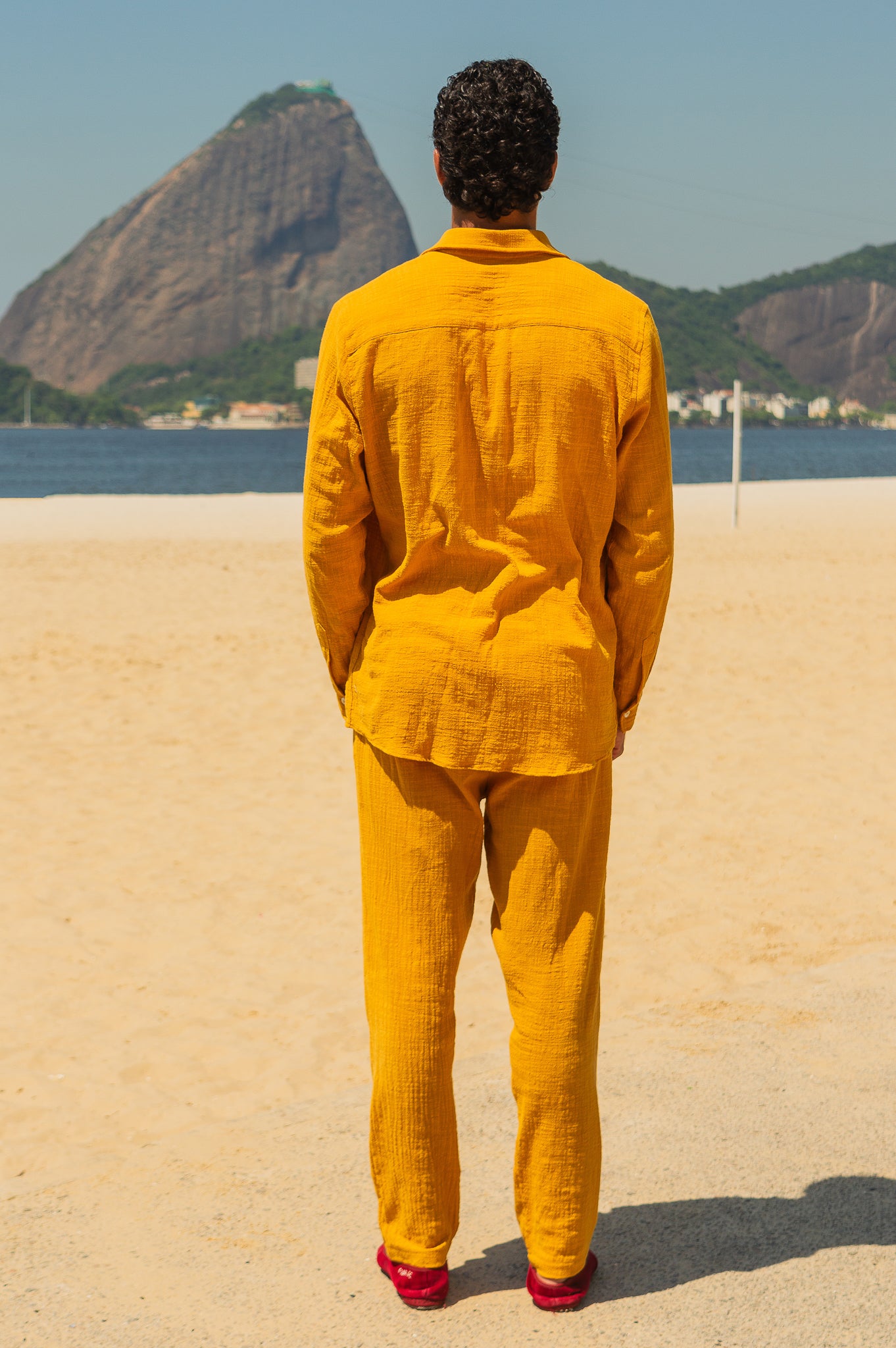 Vista traseira do full look com camisa manga longa e calça amarelo dijon unissex em modelo masculino, com cenário de praia.