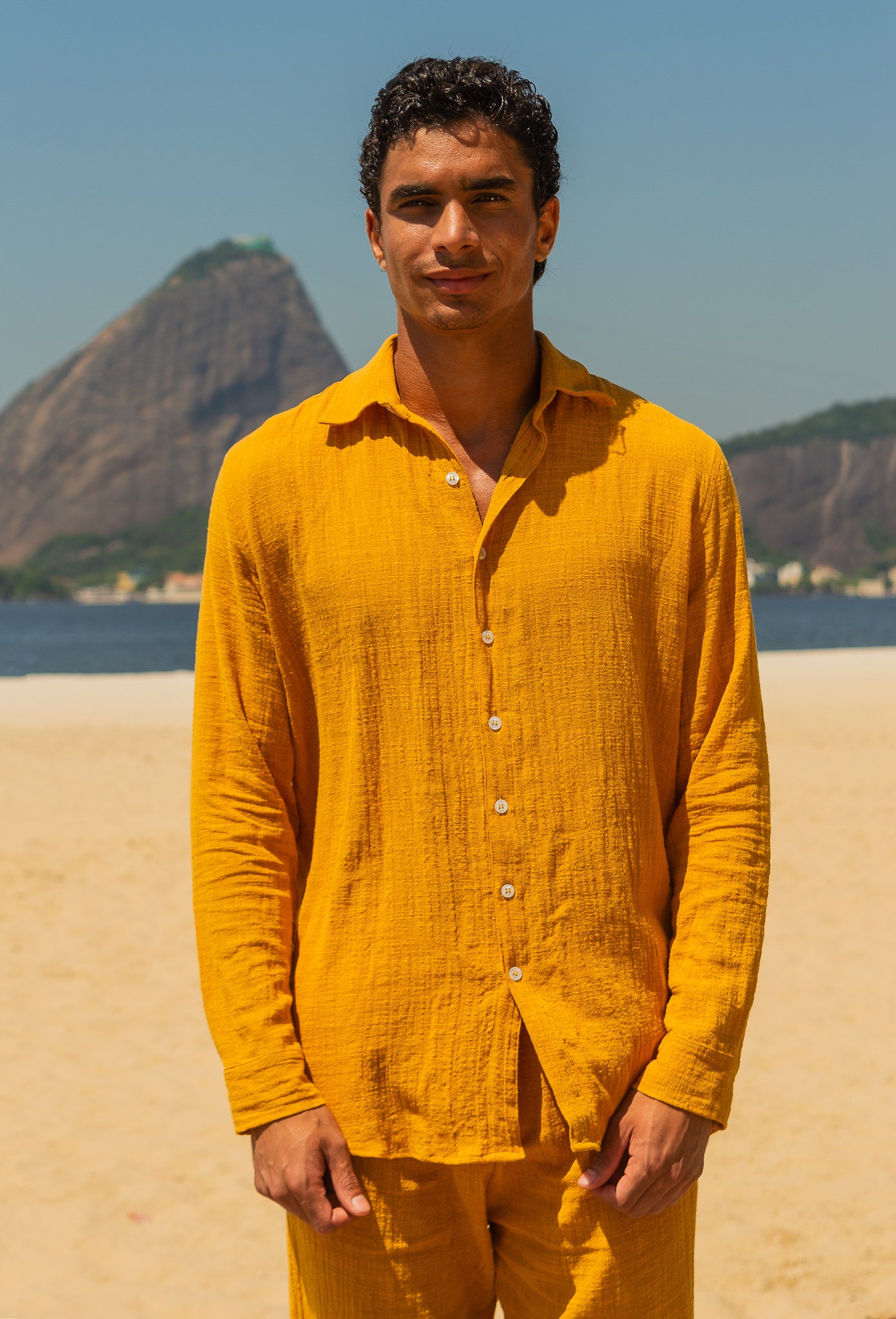Modelo masculino vestindo full look com camisa manga longa e calça amarelo dijon unissex, em pose frontal na praia.