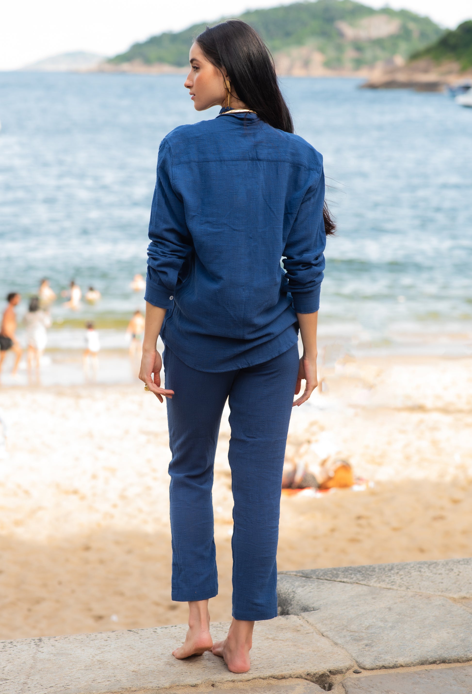 Vista traseira do full look com camisa manga longa e calça azul marinho unissex em modelo feminina, em cenário de praia.