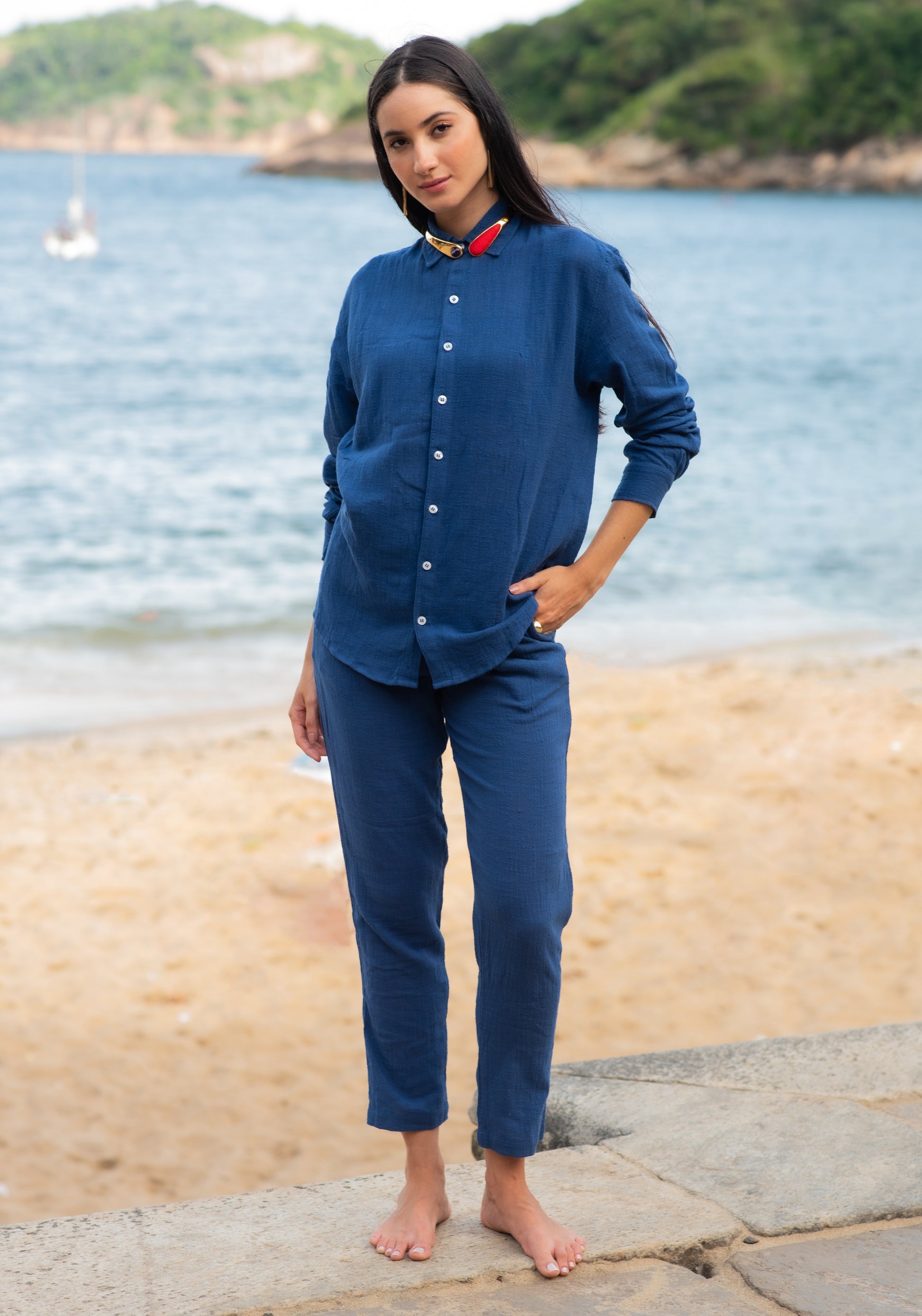 Vista frontal do full look com camisa manga longa e calça azul marinho unissex em modelo feminina, em cenário de praia.