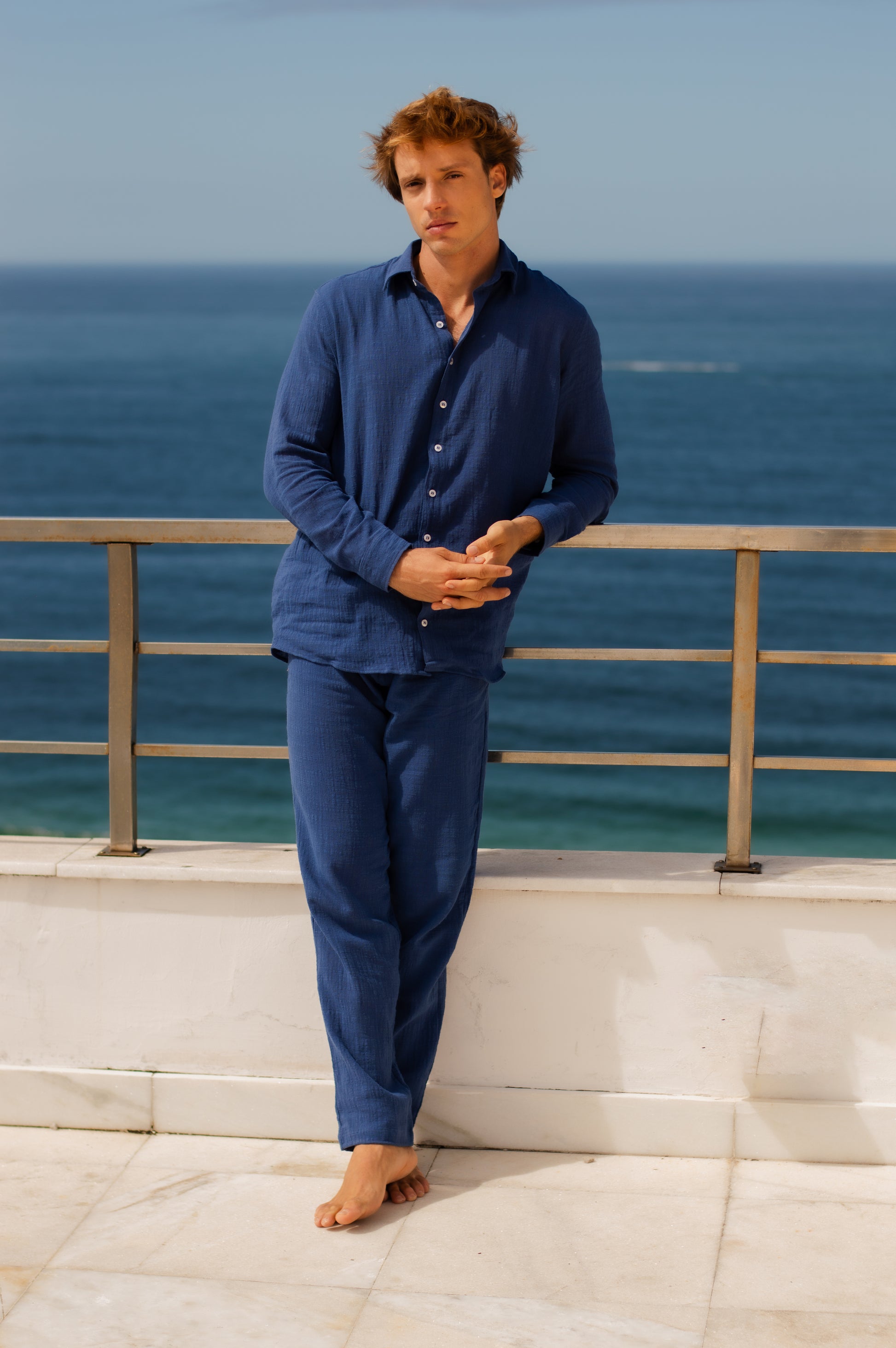 Modelo masculino vestindo full look com camisa manga longa e calça azul marinho unissex, em pose frontal, vista mar.