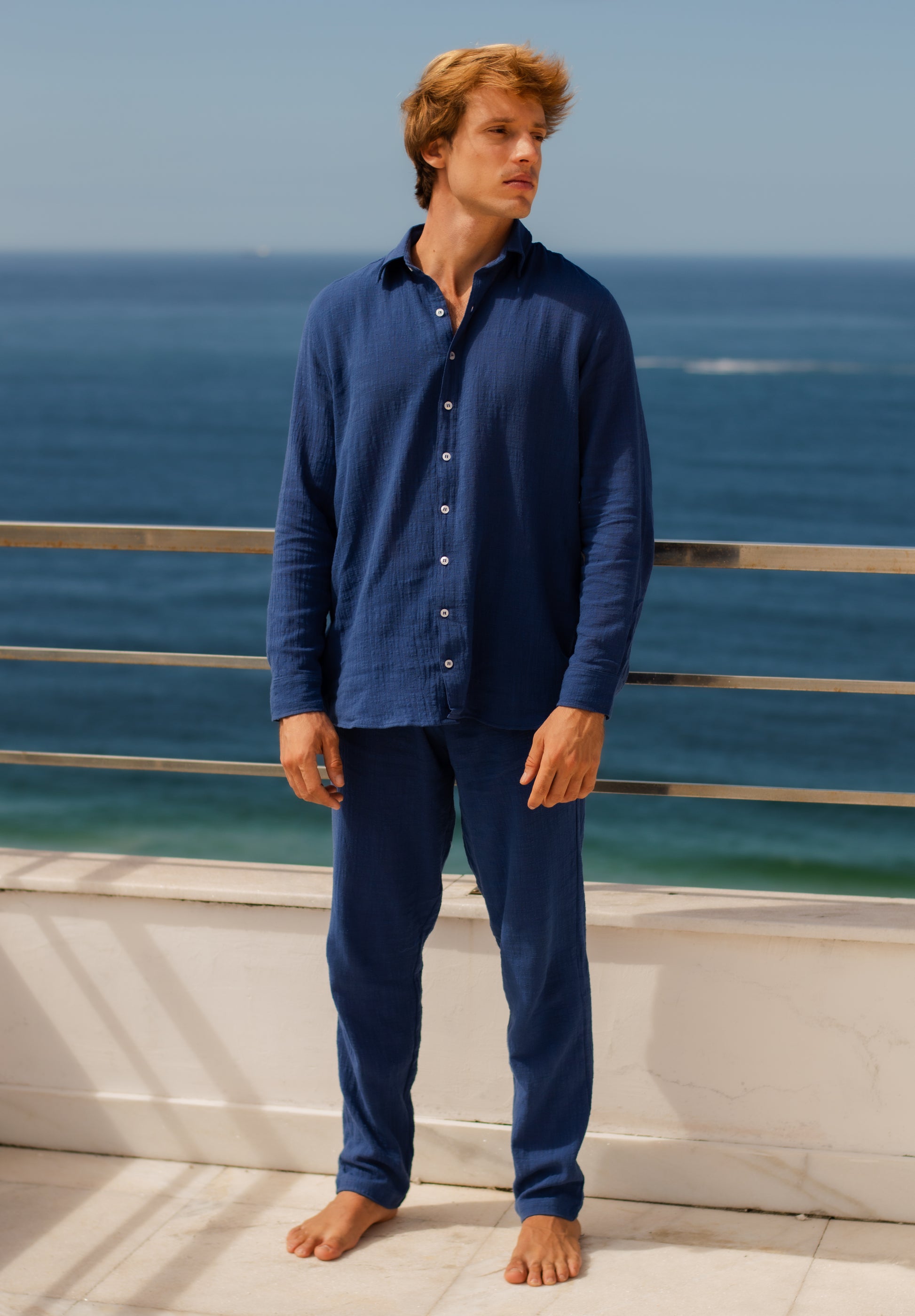 Modelo masculino vestindo full look com camisa manga longa e calça azul marinho unissex com uma vista mar.