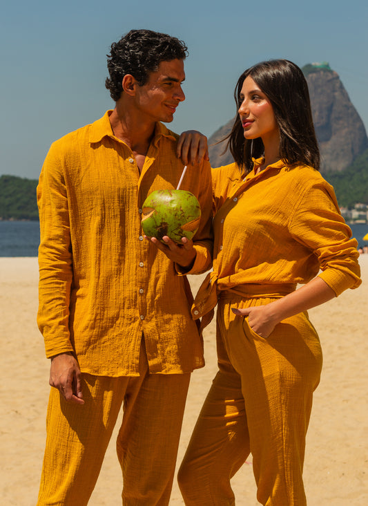 Modelo masculino e feminina vestindo full look com camisa manga longa e calça amarelo dijon unissex, segurando cocos na praia.