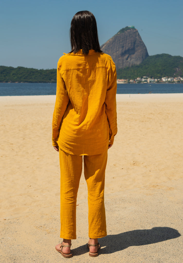 Vista traseira da camisa manga longa e calça amarelo dijon unissex em modelo feminina, com cenário de praia.