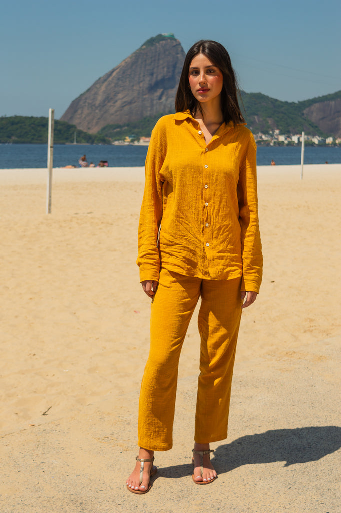 Modelo feminina vestindo full look casual descalca com camisa manga longa e calça amarelo dijon unissex, em pose frontal na praia.
