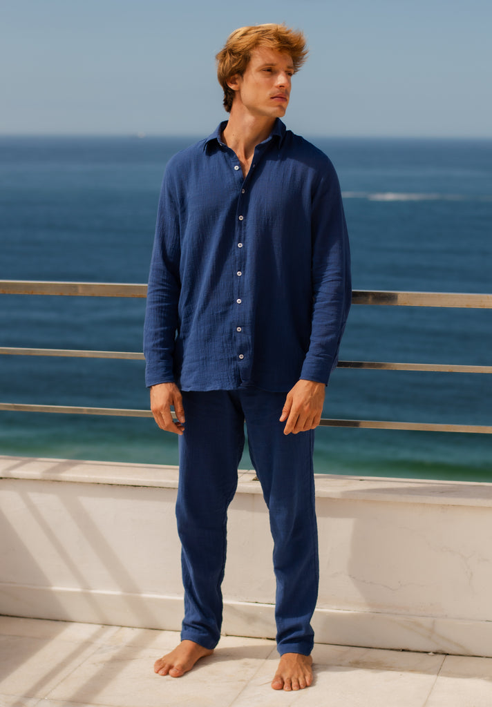 Modelo masculino vestindo full look com camisa manga longa e calça azul marinho unissex com uma vista mar.