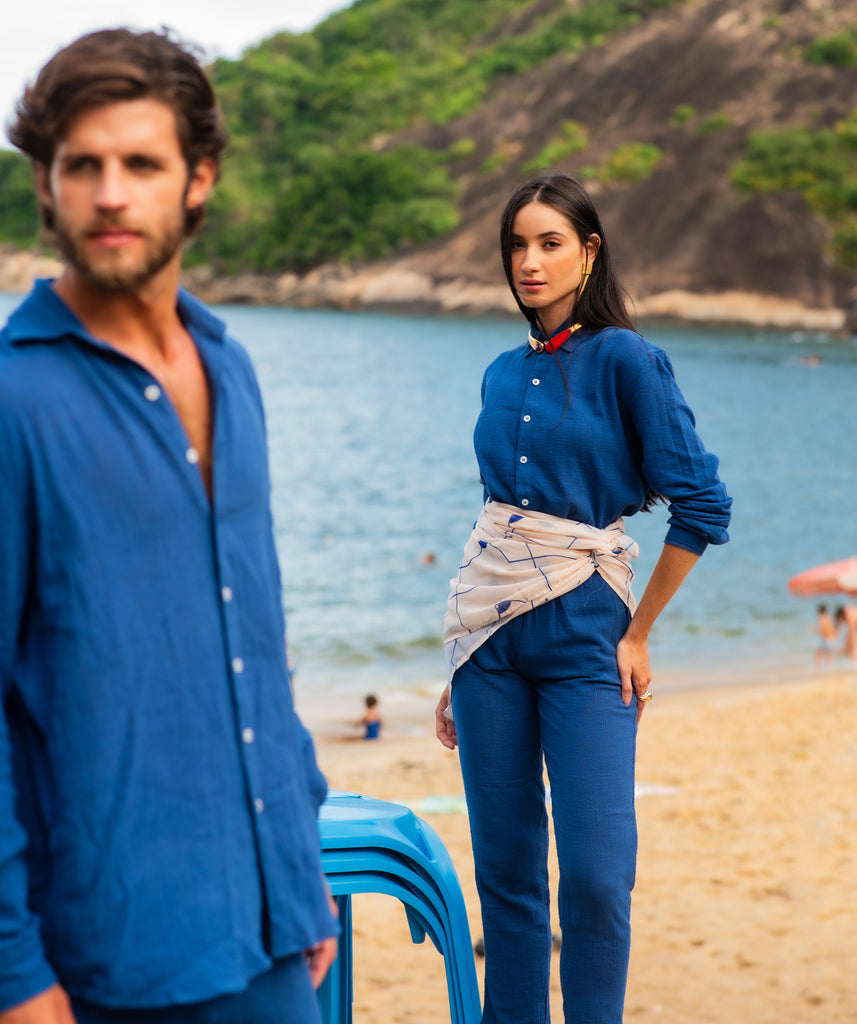Modelo masculino vestindo full look com camisa manga longa e calça azul marinho unissex, posando sentado ao ar livre.