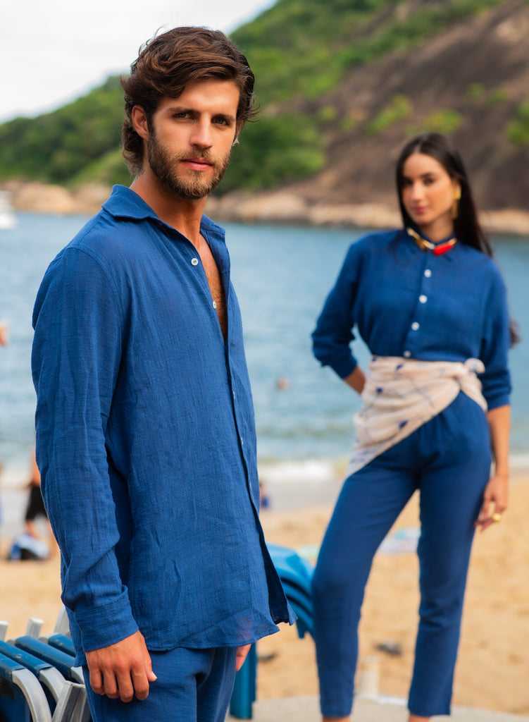 Modelo masculino e feminina vestindo full look com camisa manga longa e calça azul marinho unissex, em cenário de praia.