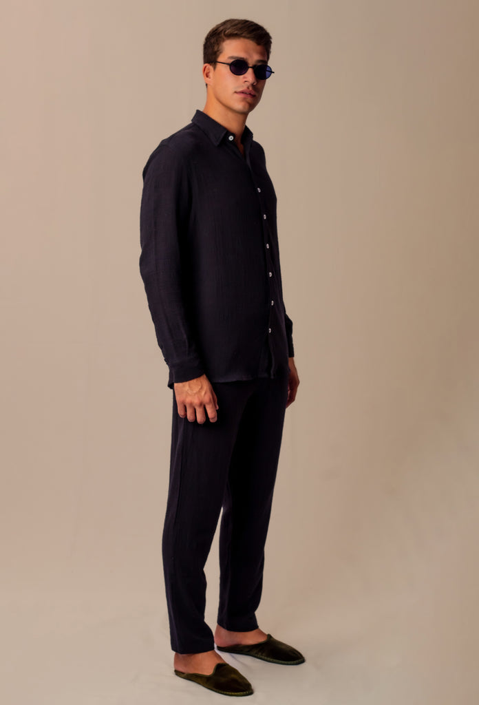 Modelo masculino usando full look com camisa manga longa e calça preta unissex, com óculos de sol, em pose lateral em fundo bege.