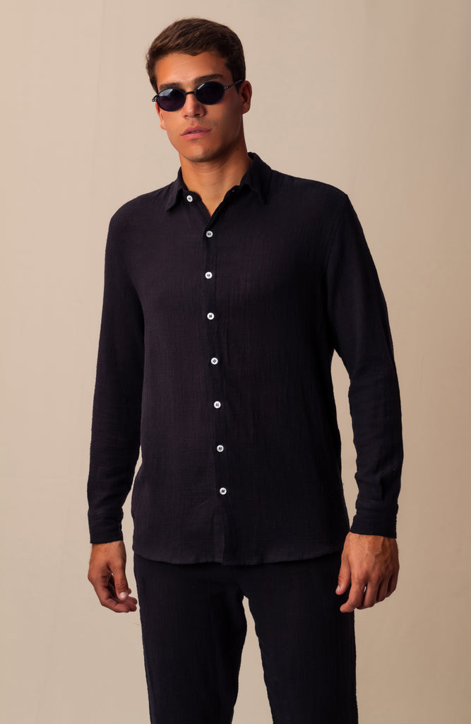 Modelo masculino usando full look com camisa manga longa e calça preta unissex, com óculos de sol, em pose lateral.